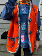 Here’s an example of an alt text for the image you sent me: “A photo of a person wearing a bright orange OMASHU jacket made of fuzzy material with a white and black braided trim, featuring a cartoon cat and mouse, with a black label on the bottom left corner, a blue sweatshirt with a cartoon mouse on it, and black pants, with long braided hair with pink and white ties at the ends, against a gray building background with a black railing.