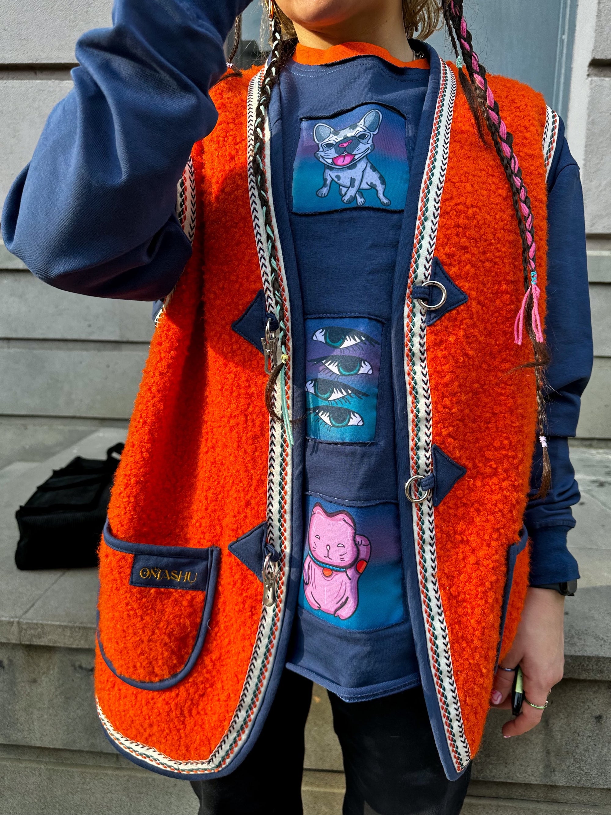Here’s an example of an alt text for the image you sent me: “A photo of a person wearing a bright orange OMASHU jacket made of fuzzy material with a white and black braided trim, featuring a cartoon cat and mouse, with a black label on the bottom left corner, a blue sweatshirt with a cartoon mouse on it, and black pants, with long braided hair with pink and white ties at the ends, against a gray building background with a black railing.