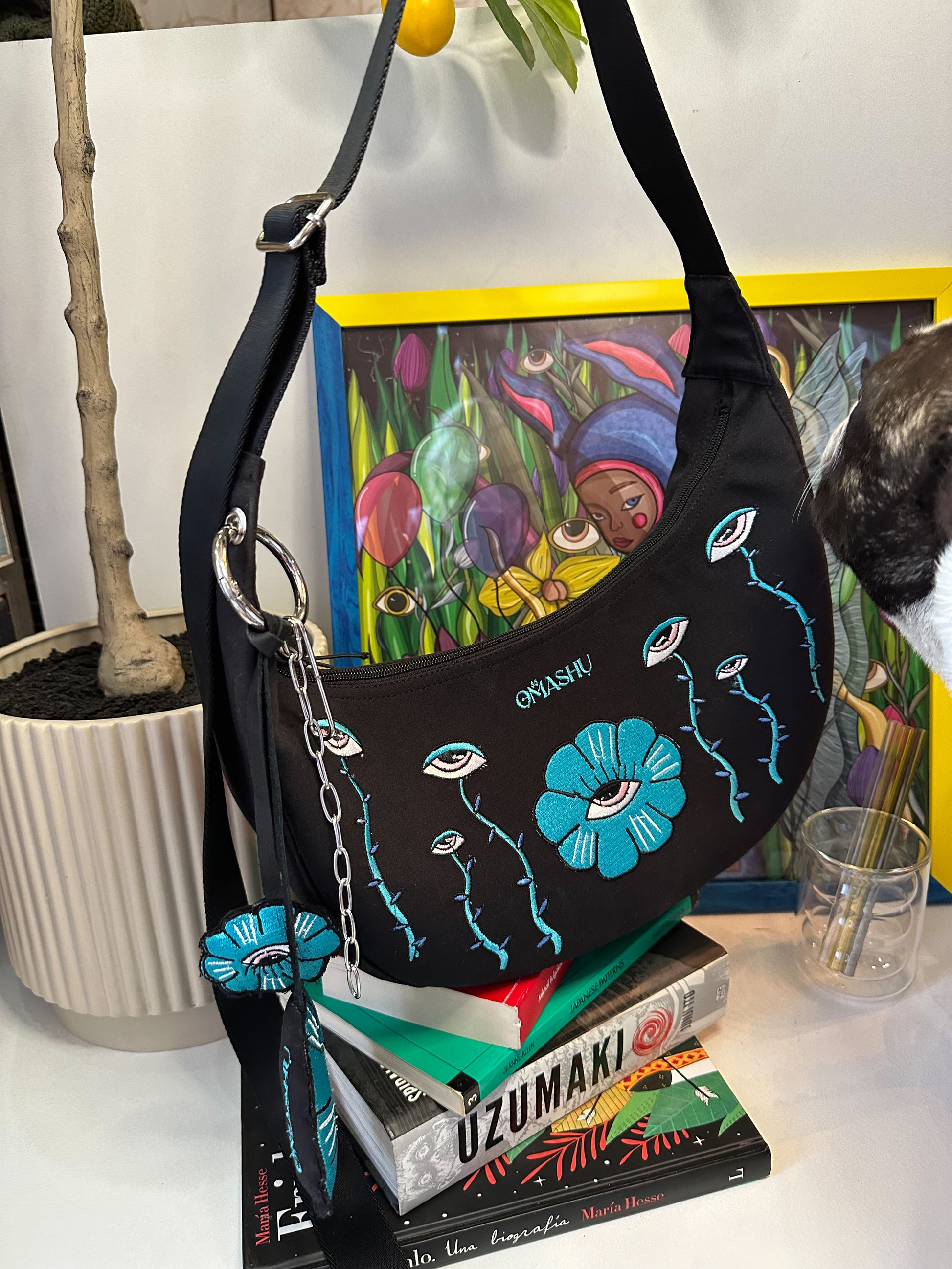 picture of OMASHU bag with teal embroidered veins and flowers with eyes. on the background there are books and pot