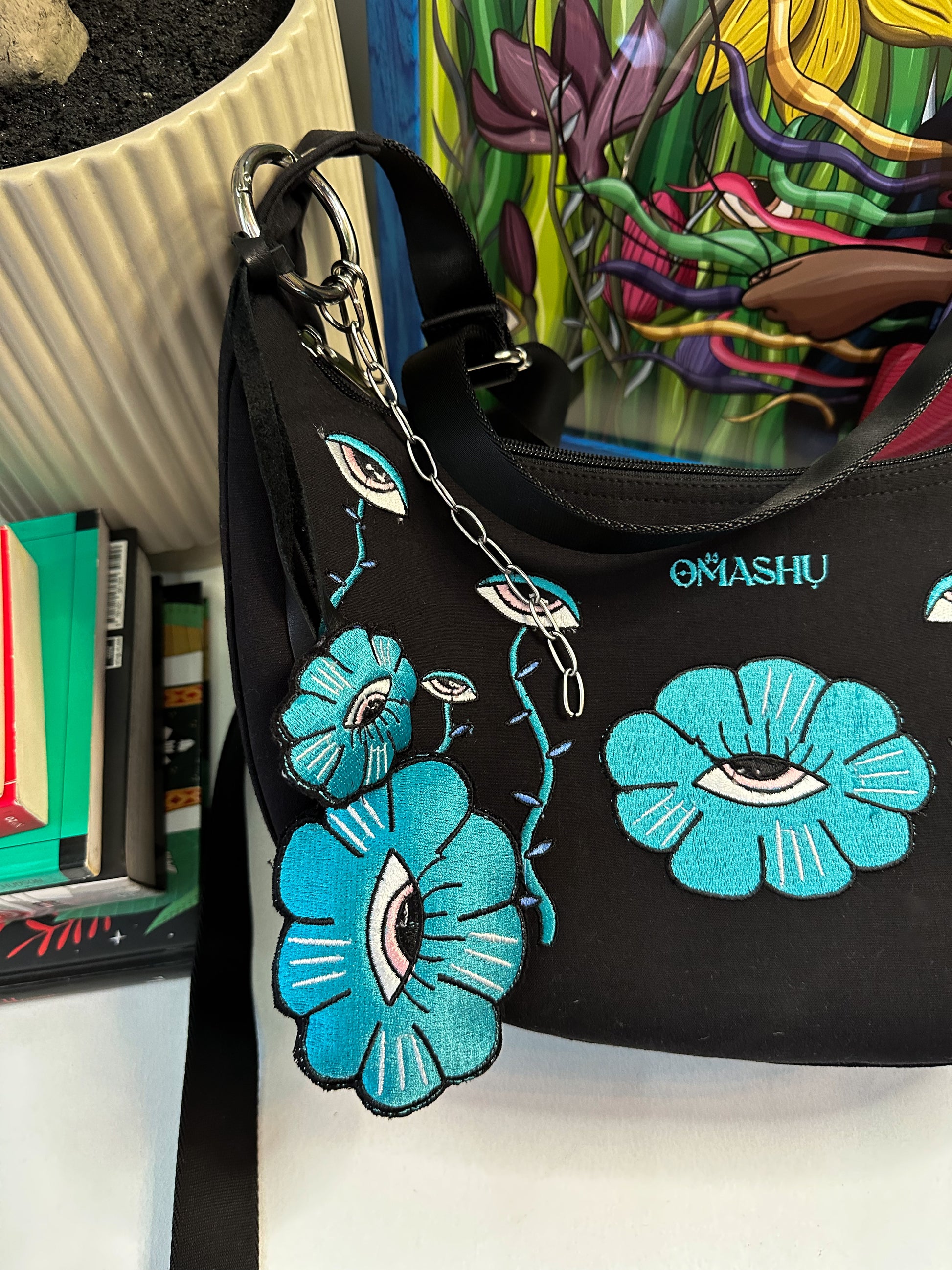 picture of a bag and key chain both has has embroidered illustration, it also has OMASHU branding on it. on the back ground there is a pot and books