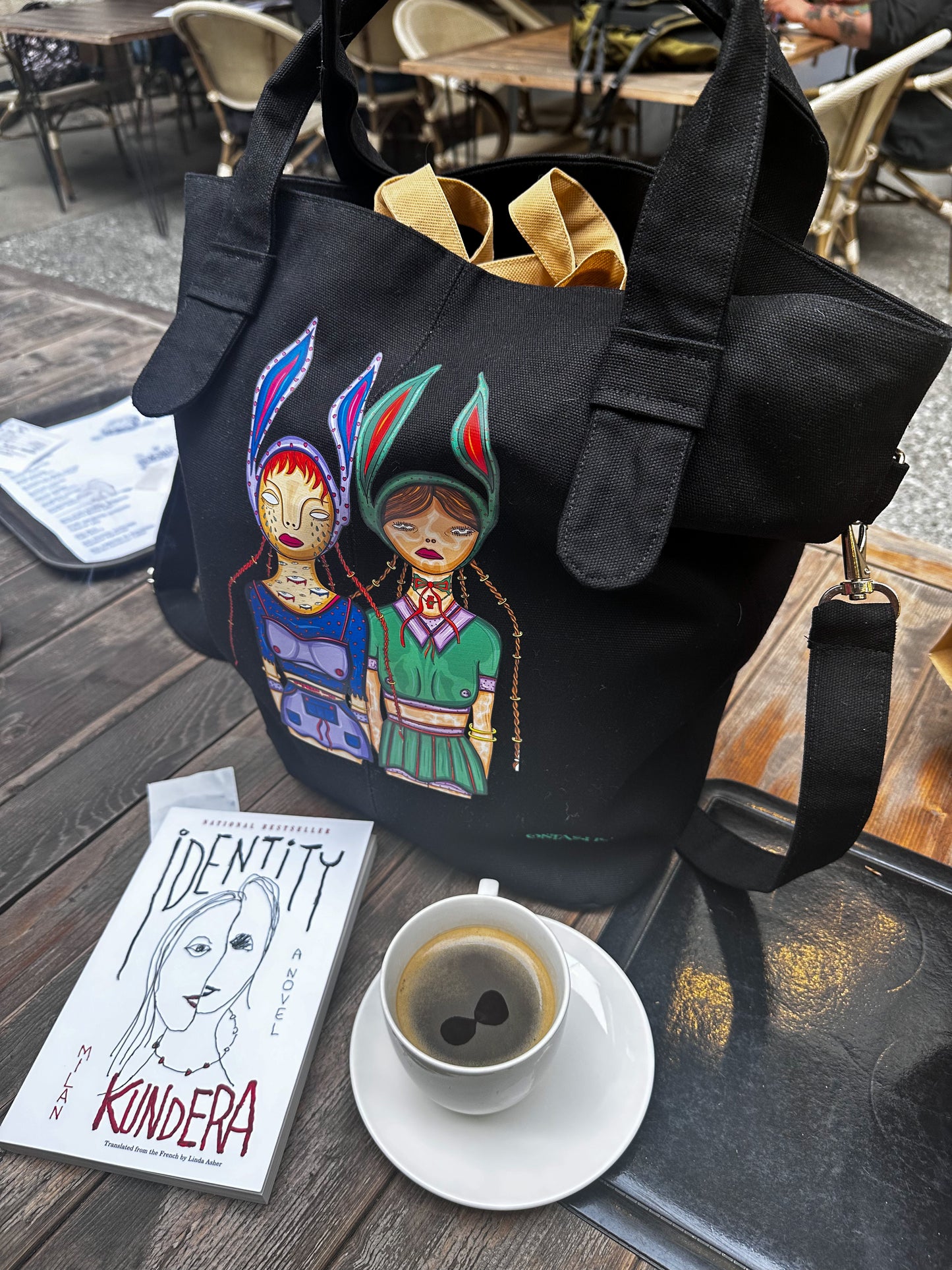 A black tote bag with a colorful design on it, a book titled ‘Identity’ by Kundera, and a cup of coffee on a table at an outdoor cafe.