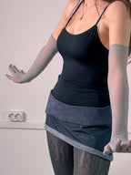 Person wearing a black tank top and gray skirt with sheer sleeves and gloves against a neutral background.