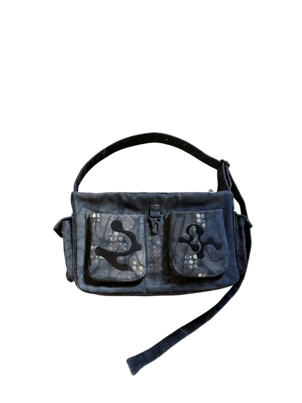 A dark blue denim messenger bag with two front pockets featuring anchor and cross appliqués, set against a light gray background with a diagonal shadow.