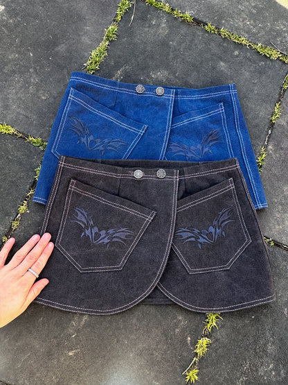 Two denim skirts, one blue and one black, both with floral embroidery on the pockets, are laid out on a gray surface with greenery in the cracks, and a person’s hand is gently touching the black skirt.