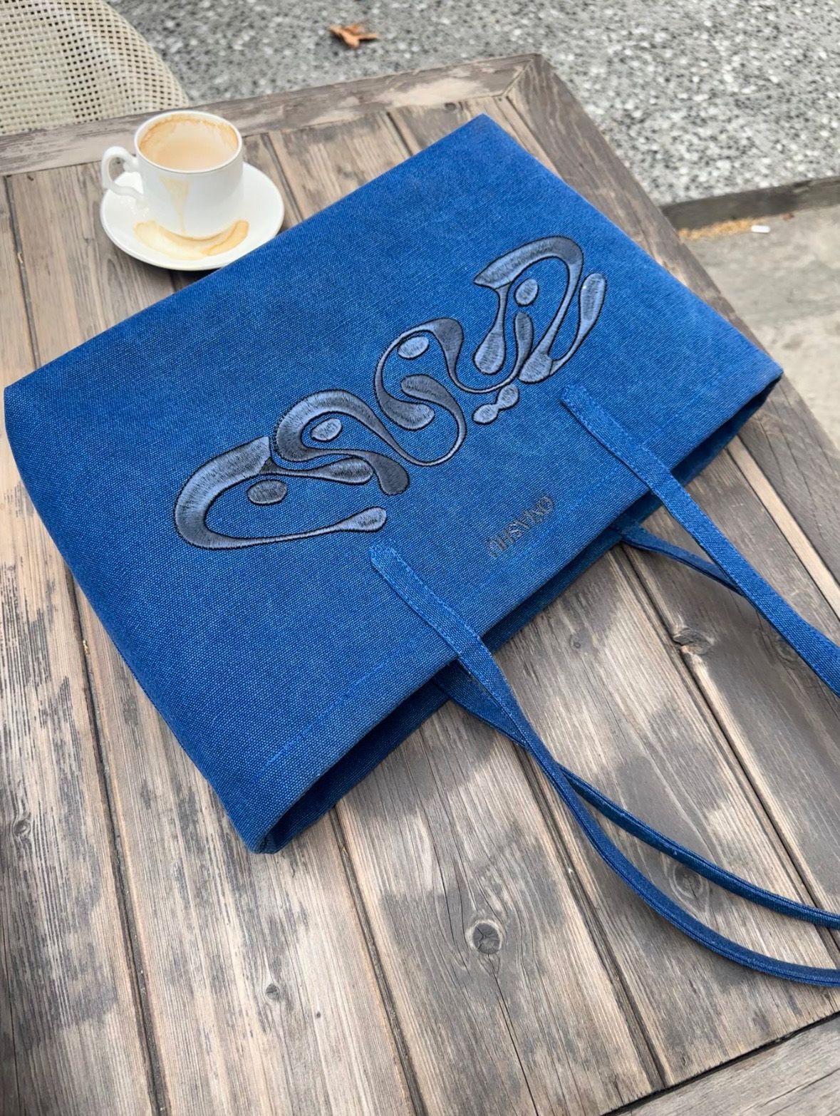 A blue tote bag with a unique design rests on a wooden surface beside a cup of coffee, suggesting a casual, artistic setting.