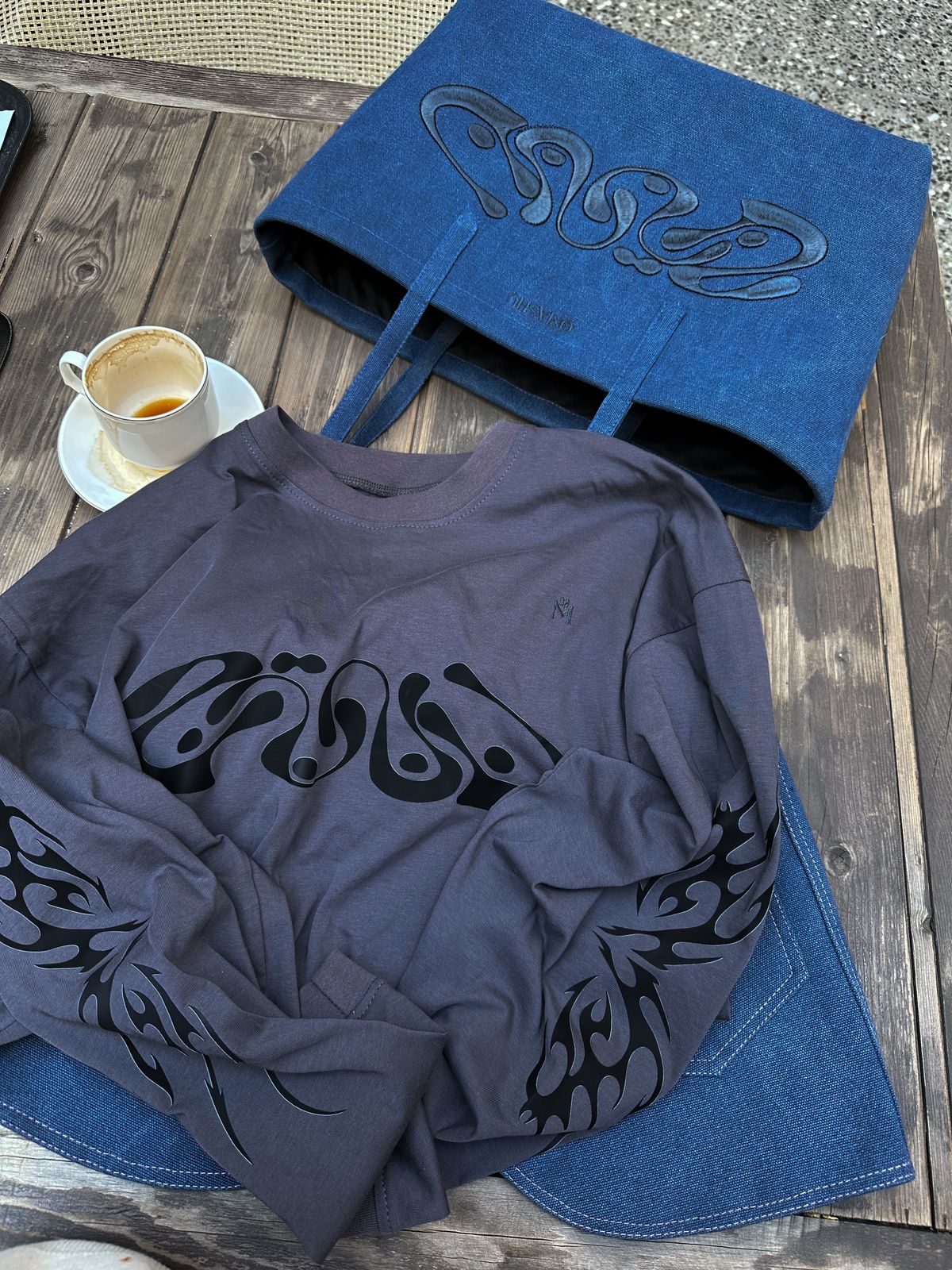 The dark grey shirt with its swirl design laid out on a table, next to a denim tote bag and a coffee cup, highlighting the intricate details of both items.