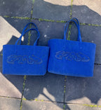 Two blue tote bags small and large with intricate designs are placed side by side on a paved surface, showcasing their symmetrical and artistic style in an outdoor setting.
