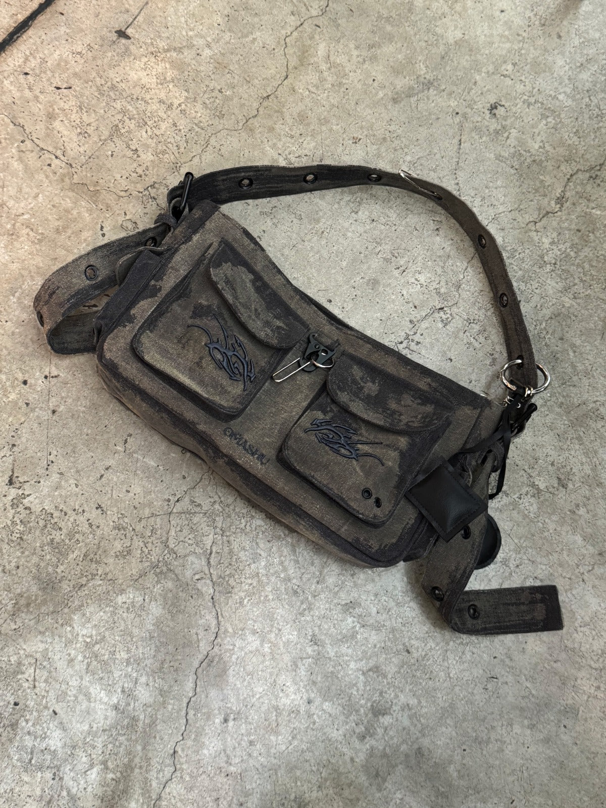 A rugged, dark gray canvas shoulder bag with a distressed, worn look, lying on a textured concrete floor. The bag features two front pockets with flap closures and an artistic black embroidery design on one pocket. The adjustable strap has metal eyelets and buckle details, giving the bag a utilitarian and vintage feel.