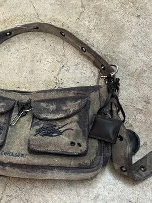 A close-up of a dark gray, rugged canvas shoulder bag resting on a textured concrete floor. The bag features two front pockets with flap closures, each with black embroidered designs. The adjustable strap includes metal eyelets, and there is a small black leather attachment on one side. The worn, distressed look of the bag gives it a vintage, utilitarian feel.