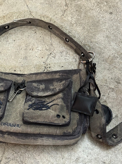 A close-up of a dark gray, rugged canvas shoulder bag resting on a textured concrete floor. The bag features two front pockets with flap closures, each with black embroidered designs. The adjustable strap includes metal eyelets, and there is a small black leather attachment on one side. The worn, distressed look of the bag gives it a vintage, utilitarian feel.