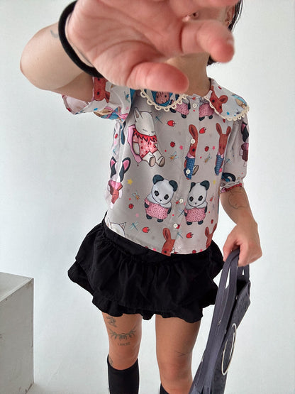 The blouse has a lace-trimmed collar and puffy sleeves, paired with a black ruffled skirt. The child's hand is partially covering the camera, adding a casual, playful feel to the image. The outfit's combination of fun prints and simple black skirt creates a stylish yet whimsical look.