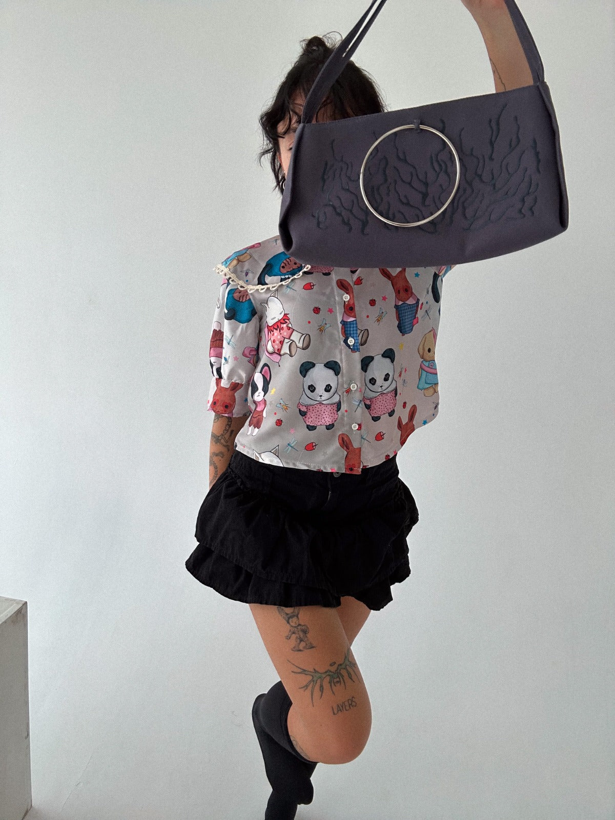 A child is wearing a colorful blouse with cartoon animal designs, including pandas and rabbits, along with a black ruffled skirt. The child is holding up a dark grey handbag with a large metal ring accent, obscuring part of their face. The playful outfit is paired with knee-high black socks, creating a fun and stylish combination. Visible tattoos on the legs add a unique detail to the overall look.