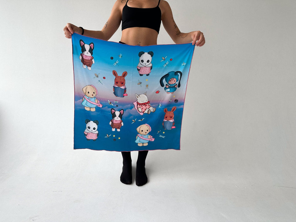 Woman holding a silk scarf with cute illustrated animal designs including a dog, panda, and rabbit, against a dreamy sky background. Fashionable and stylish, this scarf adds a whimsical flair to casual outfits. Ideal for lovers of unique, handmade accessories.
