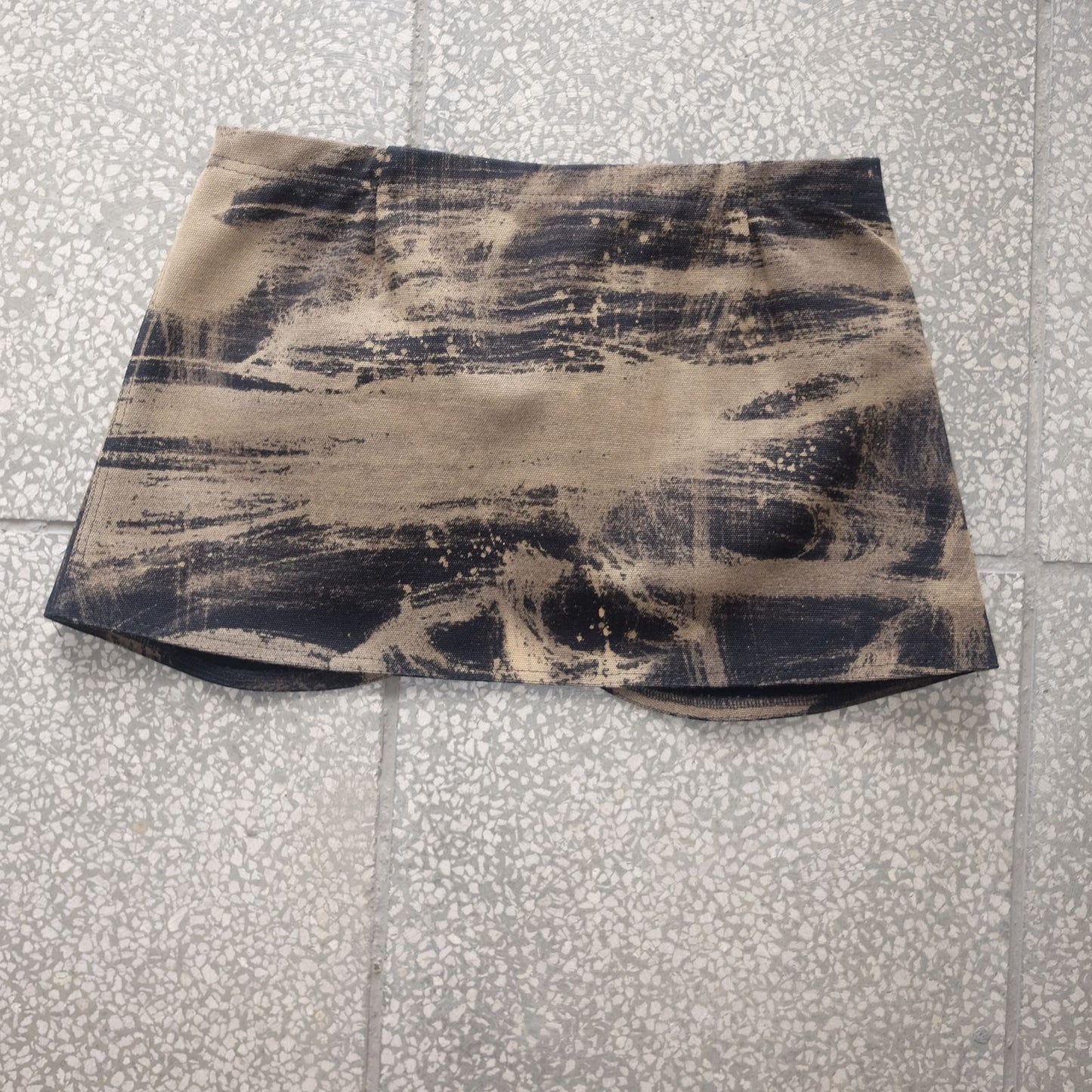 A sand-colored belt skirt with black tie-dye streaks, laid flat on a tiled surface. The abstract pattern features splattered and brushed effects, giving it a rugged, streetwear-inspired look.