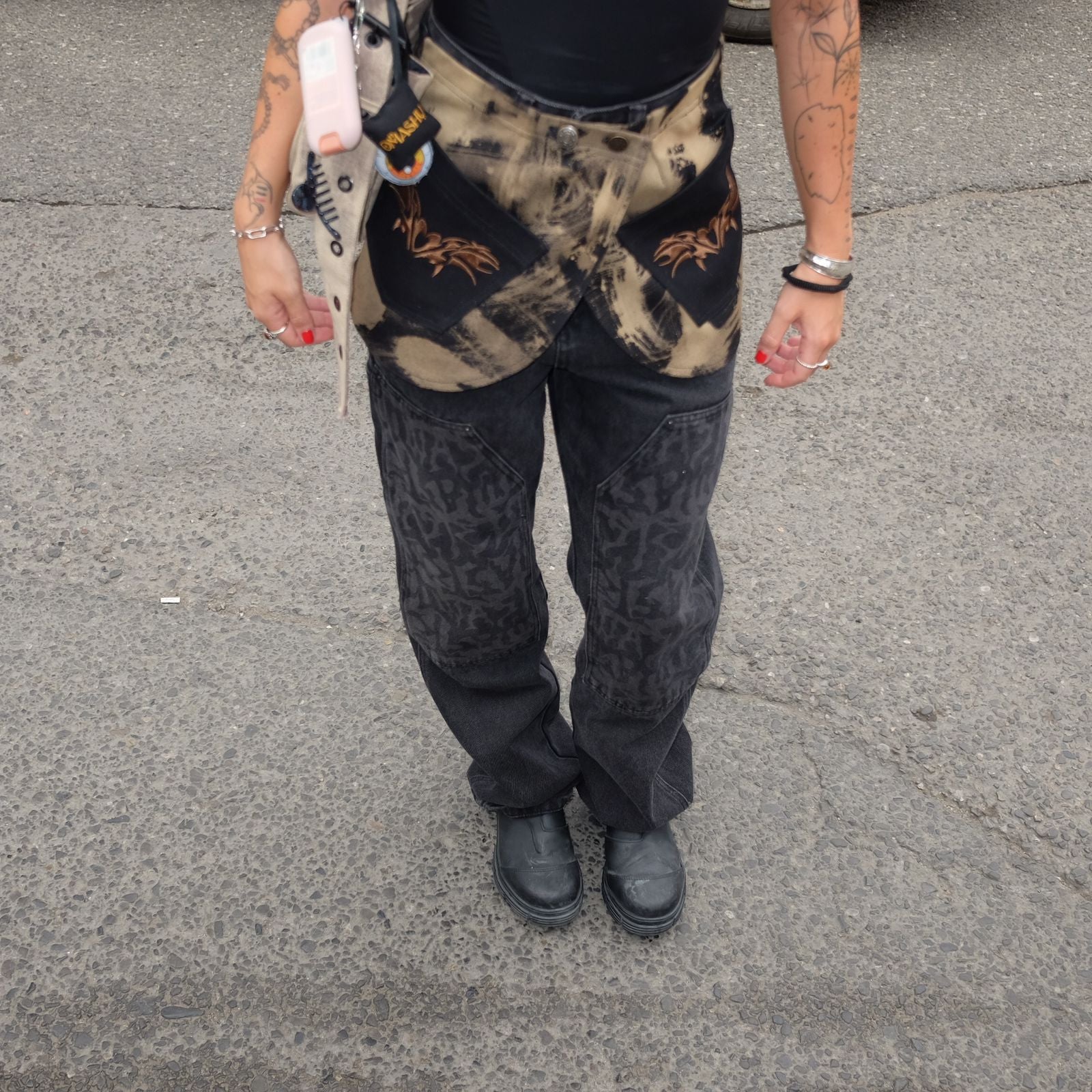 Person wearing a belt skirt in sand color with a black and tan tie-dye pattern over dark graphic pants and black chunky shoes. The outfit is accessorized with a phone case clipped to the waist, showing a casual, streetwear-inspired style.