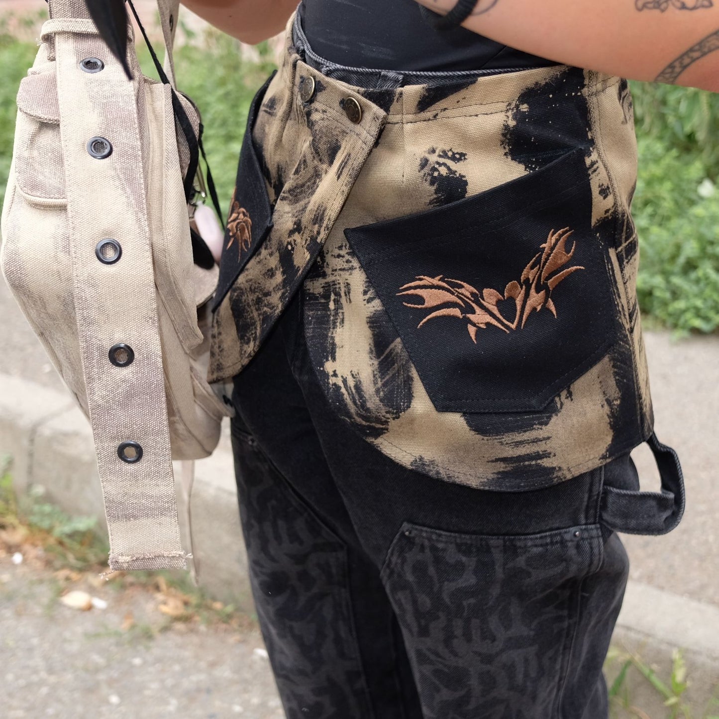 Close-up of a sand-colored belt skirt with black and tan tie-dye patterns, featuring a black pocket with an embroidered brown design. The outfit is paired with black graphic pants and a beige bag with metal grommets, highlighting the streetwear aesthetic.