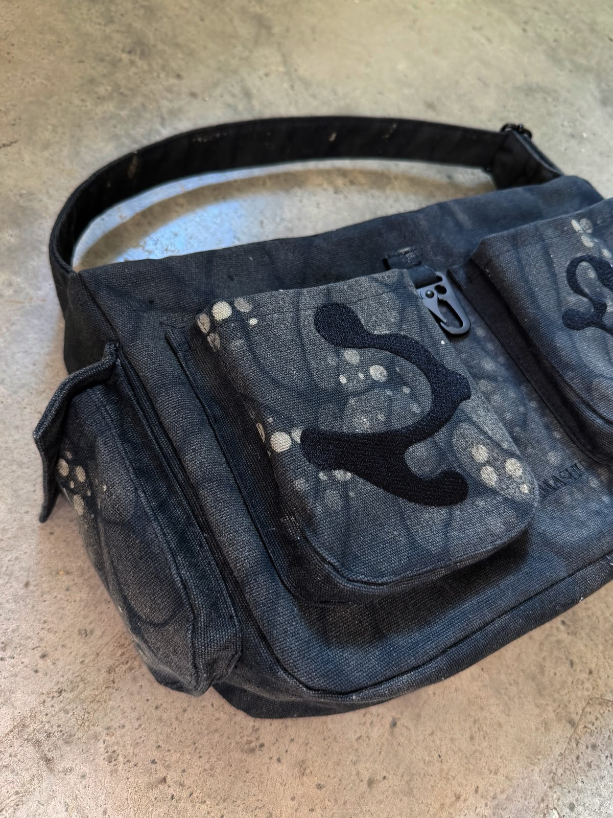 A dark gray and black canvas shoulder bag with a distressed, paint-splattered pattern and black abstract patches, resting on a concrete floor.