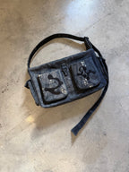 A dark denim messenger bag with black embroidered patches and silver studs, laid flat on a concrete floor.