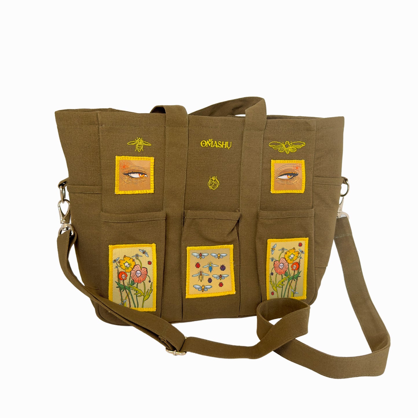 OMASHU khaki canvas bag with six pockets decorated with embroidered patches of flowers, bees, and butterflies. The top of the bag has a gold embroidered logo that reads ‘OMASHU’. The shoulder strap is adjustable and attached to the bag with gold hardware.