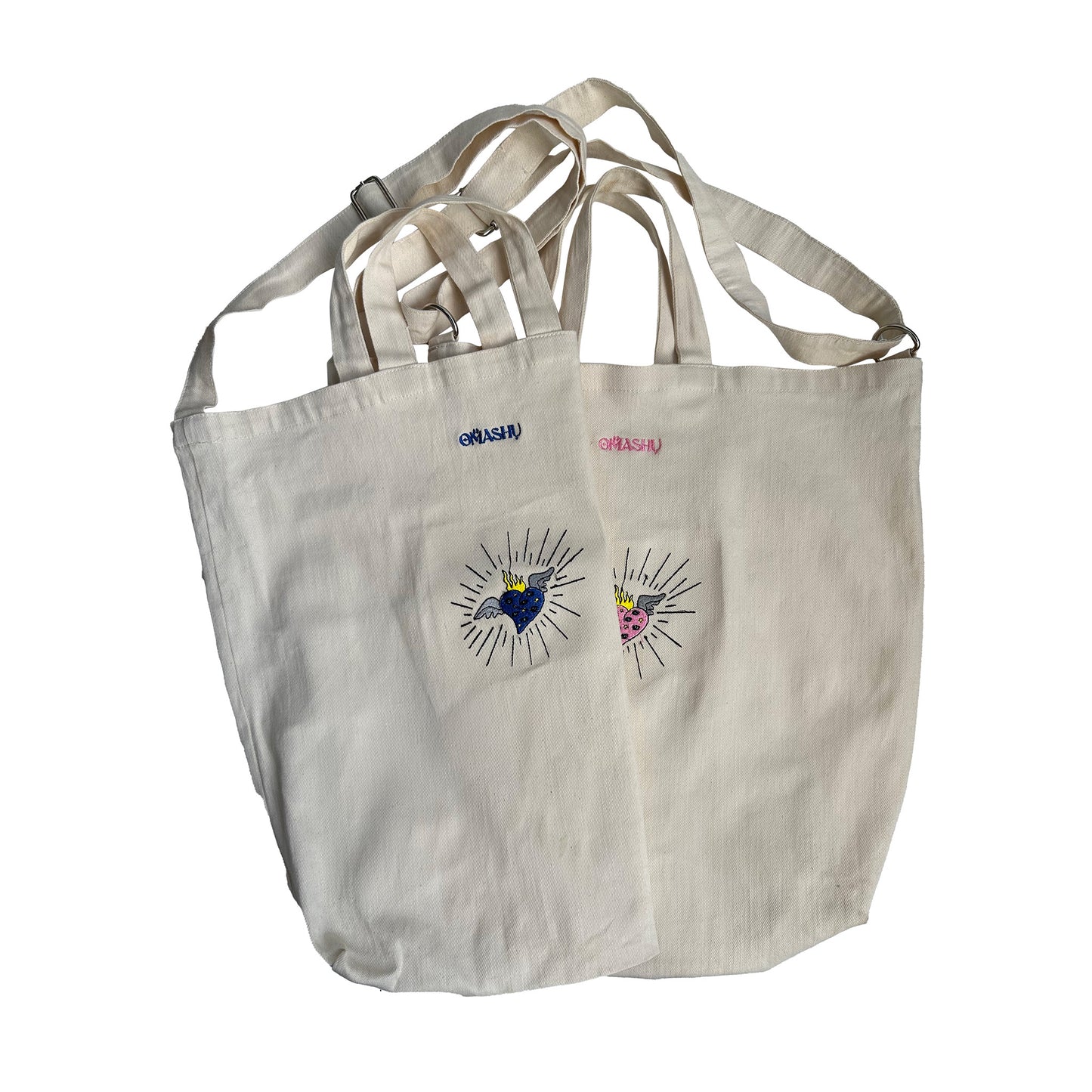 A white OMASHU tote bags made of light-colored fabric with handles, featuring two eye designs in blue and pink made up of lines and dots, creating a graphic and modern look, with the word ‘OMASHU’ written in on both bags.