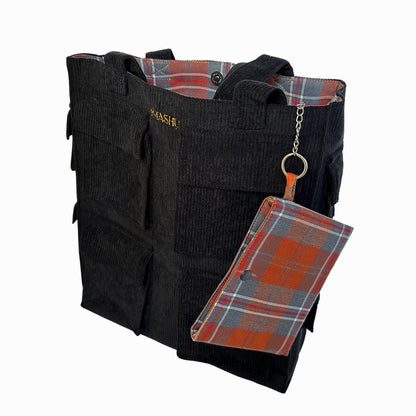 A black corduroy tote bag with a red plaid patterned pouch attached to it. The tote bag has a button closure and two handles. The pouch is attached to the tote bag with a silver chain and a key ring. The pouch has a zipper closure and a clear window on one side. The tote bag has a label on it that reads ‘OMASHU’.