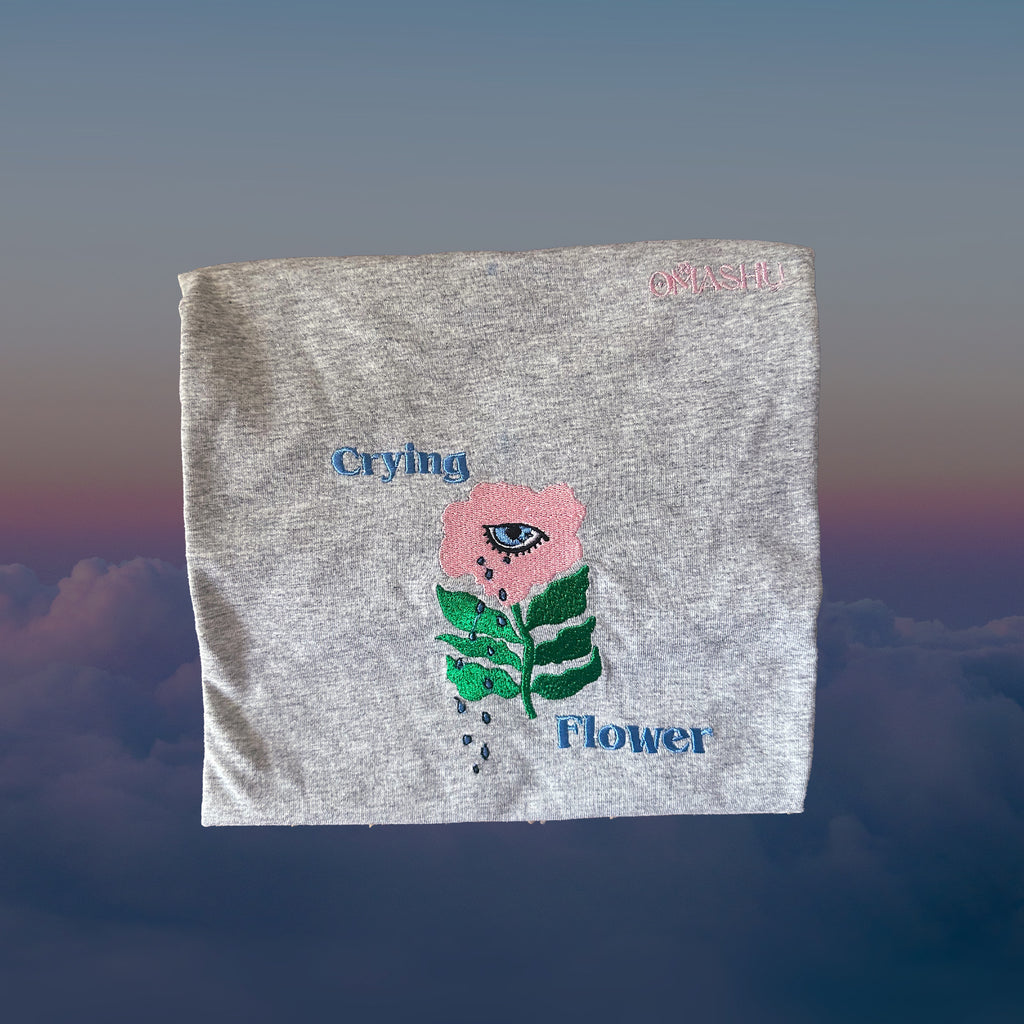 folded gray t-shirt with embroidered flower which has green stems and pink flower with eye in it. text says Crying Flower and brand is Omashu