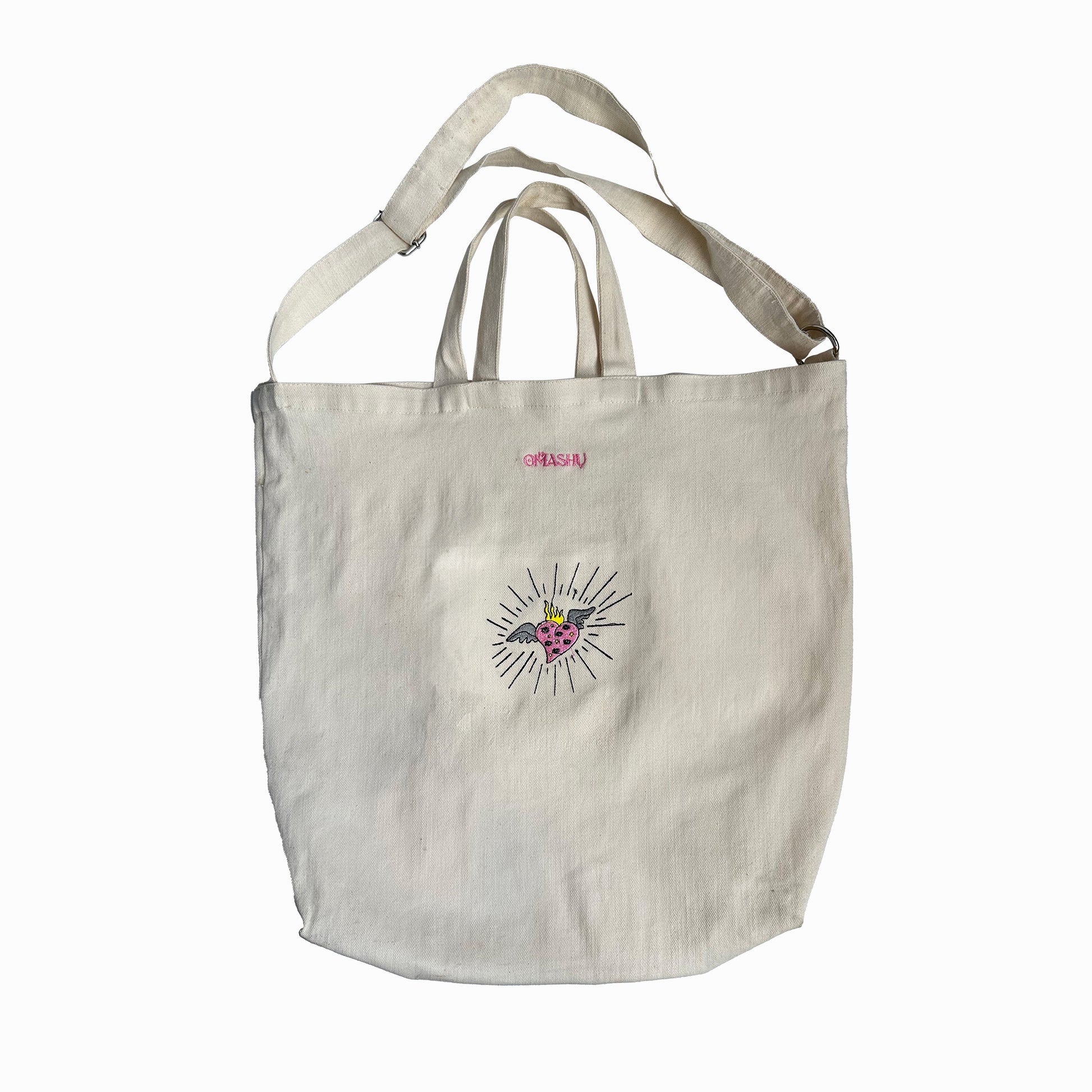 A white OMASHU tote bag with a black circle design featuring a pink heart in the center and black lines radiating outwards, with a beige strap and a small pink label on the top left corner against a white background