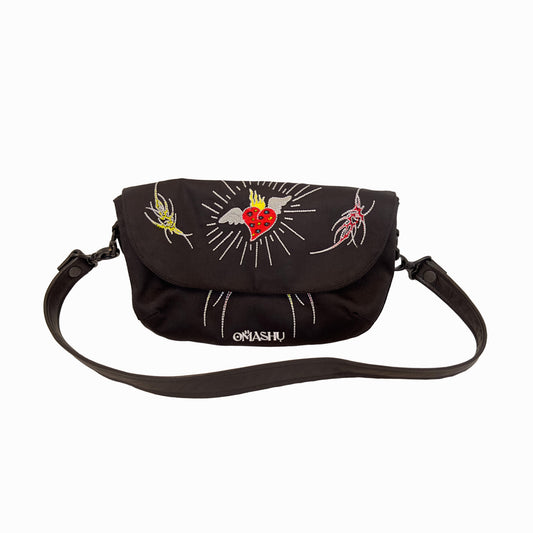 A black messenger bag with a shoulder strap. The bag has a flap with a red heart, a yellow and red tattooesque figures  The flap also has a white lines and the word ‘OMASHU’ written in white. The bag has a black shoulder strap with a black buckle.