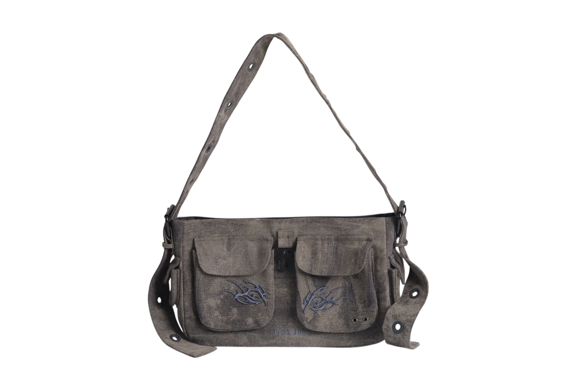 A dark gray canvas shoulder bag with a rugged, worn look, featuring two front pockets with flap closures and black embroidery designs on each pocket. The bag has an adjustable strap with metal eyelets and buckle details. A small black leather attachment hangs from one side of the bag, adding to its utilitarian aesthetic.
