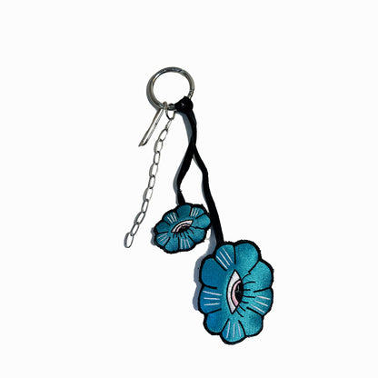 The keychain is made of a silver ring with a chain attached to it. The flowers are made of fabric and are attached to the chain with a black ribbon. The flowers are blue in color with a pink center and white details. The background is white