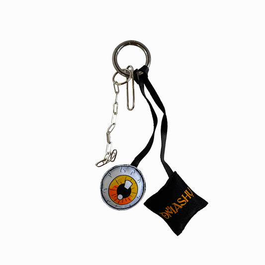 A keychain with a black ribbon and a round pendant. The pendant is a compass with a black and white face and a yellow arrow. The ribbon is black and has ‘Omashu’ written on it in orange letters. The keychain has a silver ring and chain attached to the pendant.