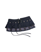 Omashu AYA Belted Layered Skirt, showing a dark navy pleated top mini skirt with silver snap buttons and an integrated corset belt with lacing, layered over a visible red, blue, and white plaid underskirt.