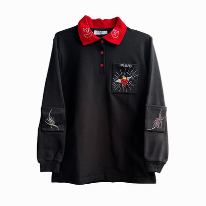 Black long sleeve polo shirt with red collar and cuffs. The shirt has a red collar with a white ‘:)’ on each side. It has a small pocket on the left chest with a white ‘OMASHU’ and a red heart embroidered on it. The shirt has a red rose embroidered on the right sleeve and a white tattooesque embroidery on the left sleeve. The shirt is displayed on a white background.