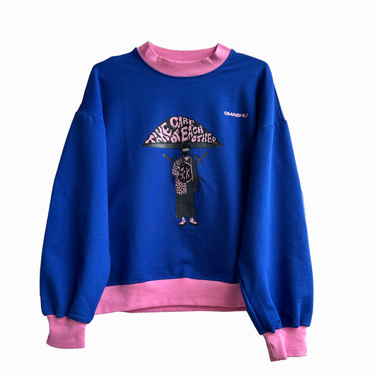 Blue and pink sweatshirt with graphic on front that reads ‘We Care For Each Other’ in black lettering. Graphic also includes illustration of person with cane and bird on shoulder. Small black logo on right shoulder that reads ‘OMASHU’.
