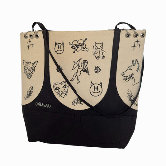 A beige and black shoulder bag with a black strap. The bag has various doodles on it such as a cat, a dog, a bee, a flower, a heart, a cross, and a person. The doodles are in black and are scattered across the beige part of the bag. The bag has two metal eyelets on the top through which the black strap is threaded. The bag has a black label on the bottom right corner that reads ‘OMASHU’.