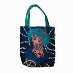 A tote bag with a graphic design on it. The tote bag is dark blue in color with a teal handle. The graphic design is of a girl with horns and a fish tail. She is holding up a peace sign with her fingers. The girl is surrounded by dark blue waves. The design is in a cartoon style. The bottom right corner of the tote bag has a goldfish in a bubble.