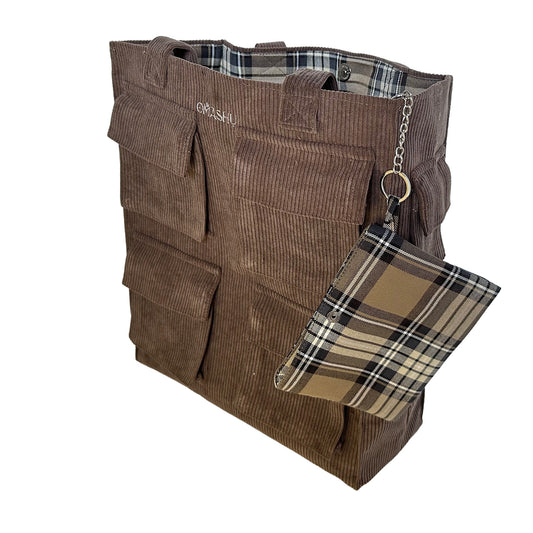 A brown corduroy tote bag with a plaid wallet attached to it. The bag has two handles and a small label on the top left corner that reads ‘OMASHU’. The wallet is attached to the bag with a silver chain and has a plaid pattern in brown, black, and white.