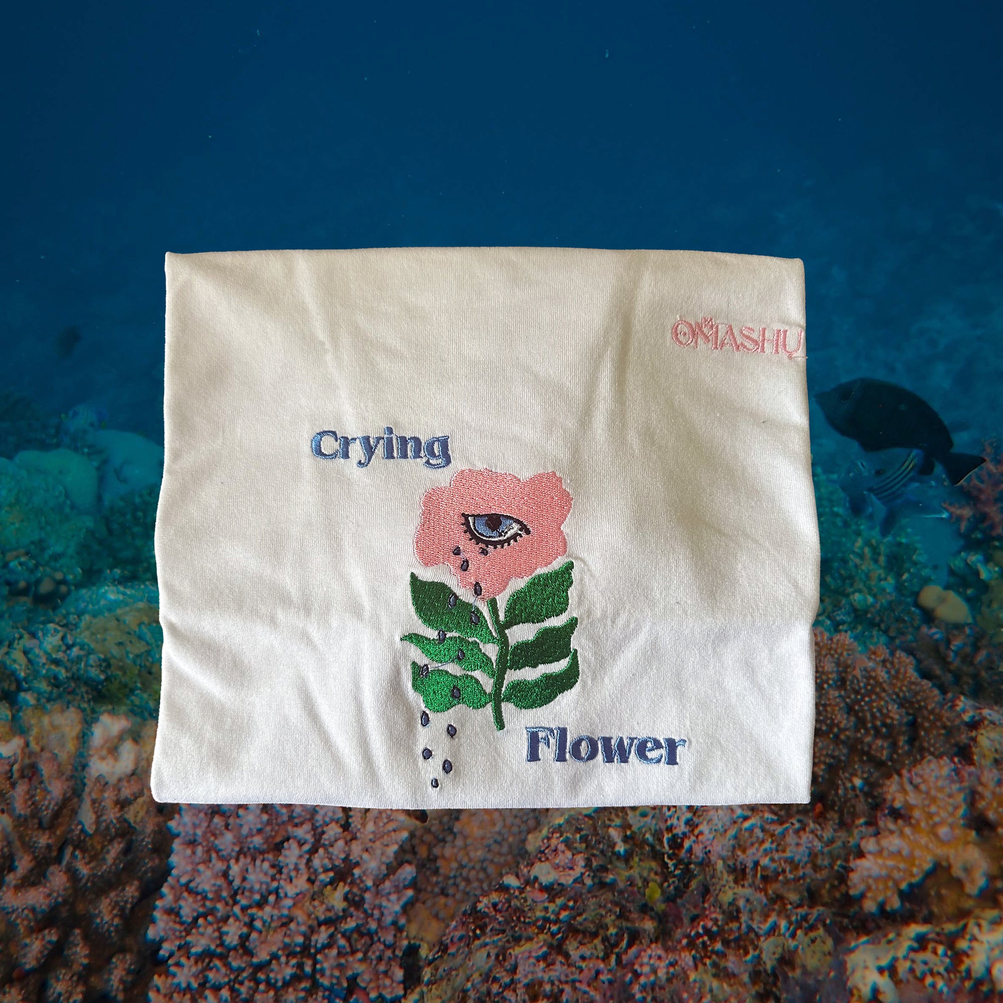 folded white t-shirt with embroidered flower which has green stems and pink flower with eye in it. text says Crying Flower and brand is Omashu